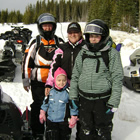 A grandmother, mother and her two daughters standing together dressed in snowmobile gear and they are surrounded by snowmobiles. 