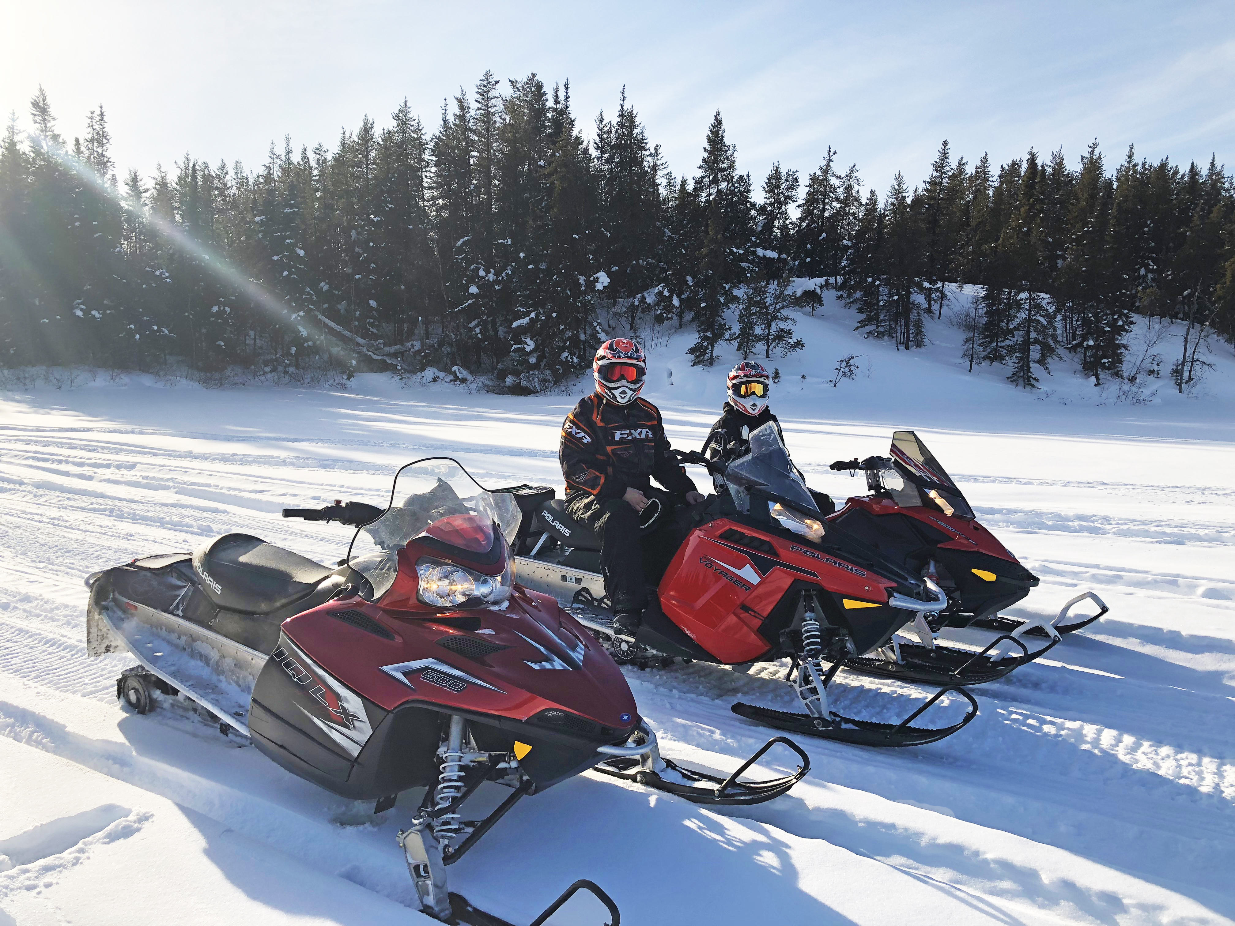 Three stationary snowmobiles, two with people sitting on them, on a flat trail with trees in the background